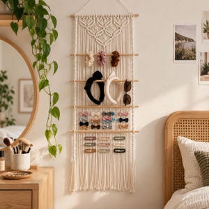 May include: A cream-colored macrame wall hanging organizer featuring five horizontal wooden dowels for displaying accessories. The organizer holds various hair scrunchies, headbands, sunglasses, small bow hair clips, and rectangular barrettes.