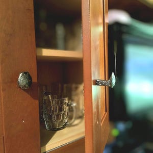 May include: A wooden cabinet door featuring a textured, silver-coloured metal handle shaped like a railway spike. The cabinet is partially open, revealing a clear glass mug with a dimpled, textured surface sitting on a wooden shelf inside the unit.