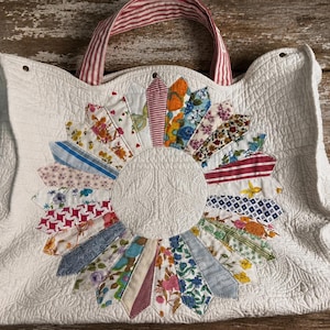 May include: A white quilted tote bag featuring a central sunburst design made of various colorful floral and patterned fabric strips. The bag has two red and white striped fabric handles and a textured, stitched surface throughout the white fabric.