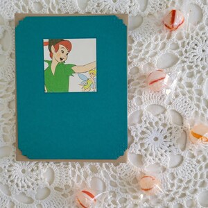 May include: A teal card with a brown border and a picture of Peter Pan and Tinkerbell. The card is on a white crocheted doily with candy.