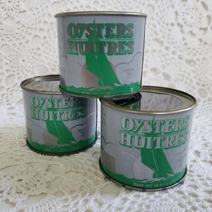 May include: Three vintage oyster cans stacked on a white lace surface. The cans are silver with green and white text and graphics. The text reads "Oysters Huîtres". Each can contains 12.5 oz.