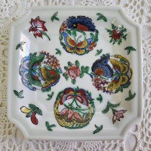 May include: A white square ceramic dish with a decorative design of colorful butterflies and flowers. The dish has a slightly raised edge and a scalloped border.