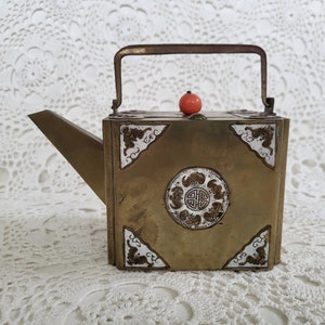 May include: A vintage brass teapot with a rectangular body and a long spout. The teapot features decorative panels with Asian-inspired designs and a small red knob on the lid. It has a handle for carrying.