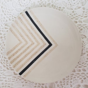 May include: A white ceramic plate with a black and beige geometric design. The design features a series of thin, horizontal lines that create a chevron pattern.