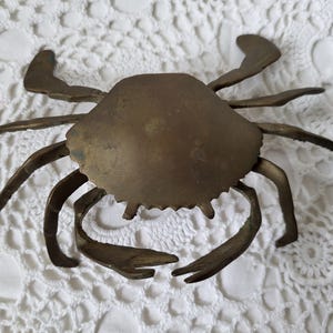 May include: A brass crab figurine featuring a smooth, rounded central shell and eight articulated legs extending outward. The metal surface displays a muted, aged golden-brown patina with subtle variations in tone across the entire structure.