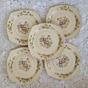 May include: Set of five square-shaped ceramic plates with a cream background and gold trim. Each plate features a floral design with purple, yellow, and orange flowers.