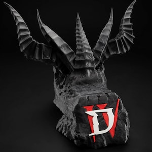 May include: A dark gray, textured sculpture featuring a central crown-like structure with multiple sharp, pointed horns extending outward. The front face of the base displays a prominent white letter D overlaid on a vertical red line graphic.