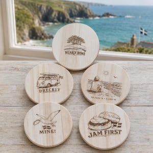 May include: Five round light-colored wooden coasters featuring laser-engraved illustrations. Designs include a tree, a camper van with the text DRECKLY, a person on a beach with coordinates, a seagull with the text MINE!, and a scone with the text JAM FIRST.