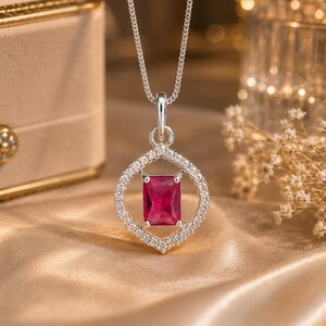 May include: A silver pendant necklace featuring a rectangular, deep pink gemstone set in the center of a marquise-shaped frame encrusted with small, clear, sparkling stones. The pendant hangs from a delicate silver chain with a fine link design.