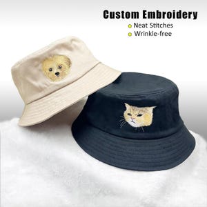 May include: A beige bucket hat with an embroidered dog face and a black bucket hat with an embroidered cat face. Text reads: Custom Embroidery, Neat Stitches, Wrinkle-free. Both hats feature a wide brim and a rounded crown design.