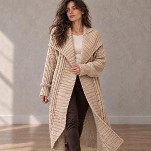 May include: A long, beige, open-front knit cardigan featuring a wide shawl collar, ribbed texture, and long sleeves with cuffed wrists. The garment is worn over a white top and dark trousers, showcasing a heavy, chunky knit construction throughout.