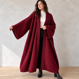 Puede incluir: Un cárdigan largo hasta el suelo de color burdeos con frente abierto, textura de punto grueso, mangas anchas estilo kimono y un borde acanalado a lo largo de la abertura. La prenda se lleva sobre una blusa blanca de cuello alto y pantalón negro.