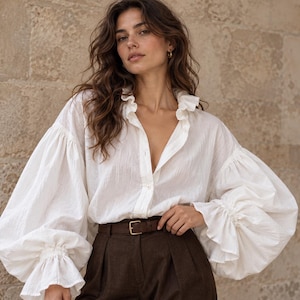 Peut inclure: Une chemise blanche à manches longues boutonnée avec des manches bouffantes volumineuses, des poignets à volants et un col à volants, portée avec un pantalon marron foncé maintenu par une ceinture en cuir marron à boucle rectangulaire dorée.