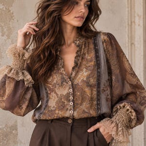 Puede incluir: Una blusa de manga larga en tonos marrón y beige con estampado de cachemira, cierre frontal de botones y cuello en V con volantes. La prenda incluye paneles de tela semitransparente y puños anchos fruncidos con ribete de encaje beige.
