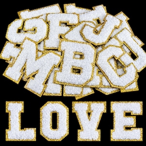 Può includere: Una collezione di toppe a forma di lettera in ciniglia bianca in stile varsity con bordi glitterati dorati, disposte in un mucchio con le lettere L, O, V e E visualizzate in una riga in basso su uno sfondo nero uniforme.