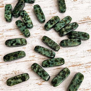 May include: A collection of rectangular, polished beads with rounded edges, featuring a mottled pattern of dark forest green and black spots throughout the smooth, glossy surface of each individual stone piece.