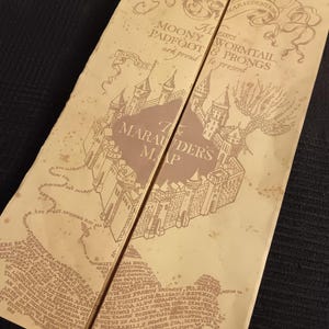 Op de afbeelding: Een beige perkamentachtige Marauder's Map met bruine inktillustraties van een kasteel, kronkelende paden en tekst waaronder "Messrs. Moony, Padfoot, Wormtail, & Prongs are proud to present The Marauder's Map" met decoratieve krulmotieven.