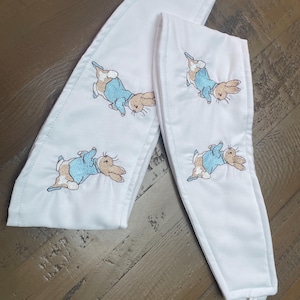 May include: Two white fabric straps with embroidered Peter Rabbit images. The straps have a loop at the top and a tie at the bottom.
