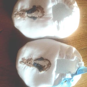 May include: A pair of white baby booties with blue ribbon ties. Each bootie features an embroidered brown rabbit with a blue bow tie.