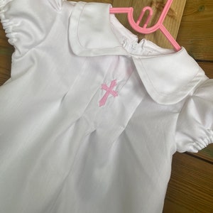 Puede incluir: Batista blanca para bebé con una cruz bordada en rosa en la parte delantera. La bata tiene un cuello Peter Pan y mangas cortas.