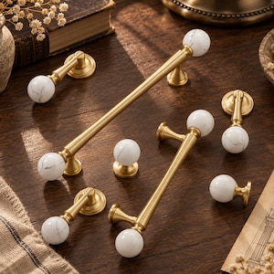 May include: A collection of cabinet hardware including two long bar pulls, two drop pulls, and two round knobs. Each piece features a brushed gold metal finish paired with white marble spheres that have subtle grey veining throughout the stone.
