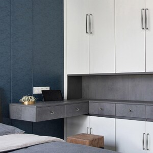 May include: A wall-mounted desk unit featuring a dark gray stone-textured surface with two drawers, positioned below white cabinetry with vertical handles. A black tablet and a small potted plant sit on the desk, set against a dark blue wall.