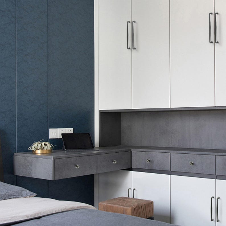 May include: A wall-mounted desk unit featuring a dark gray stone-textured surface with two drawers, positioned below white cabinetry with vertical handles. A black tablet and a small potted plant sit on the desk, set against a dark blue wall.