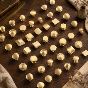 May include: A collection of various brass cabinet knobs in multiple shapes including round, square, hexagonal, and faceted designs. These metallic hardware pieces feature a smooth, brushed gold finish and are arranged in a grid pattern.