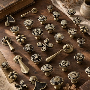 May include: A collection of antique brass cabinet knobs and drawer pulls in various shapes, including floral, circular, and fan designs. The hardware features intricate embossed patterns and a weathered metallic finish, arranged on a wooden surface.