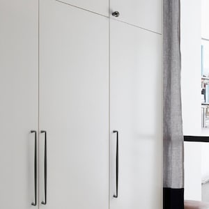 May include: A white wardrobe cabinet featuring three tall, flat-panel doors with long, slender, dark metal vertical handles and a single small, round, dark metal knob on the upper right door panel, providing a clean and functional storage solution.