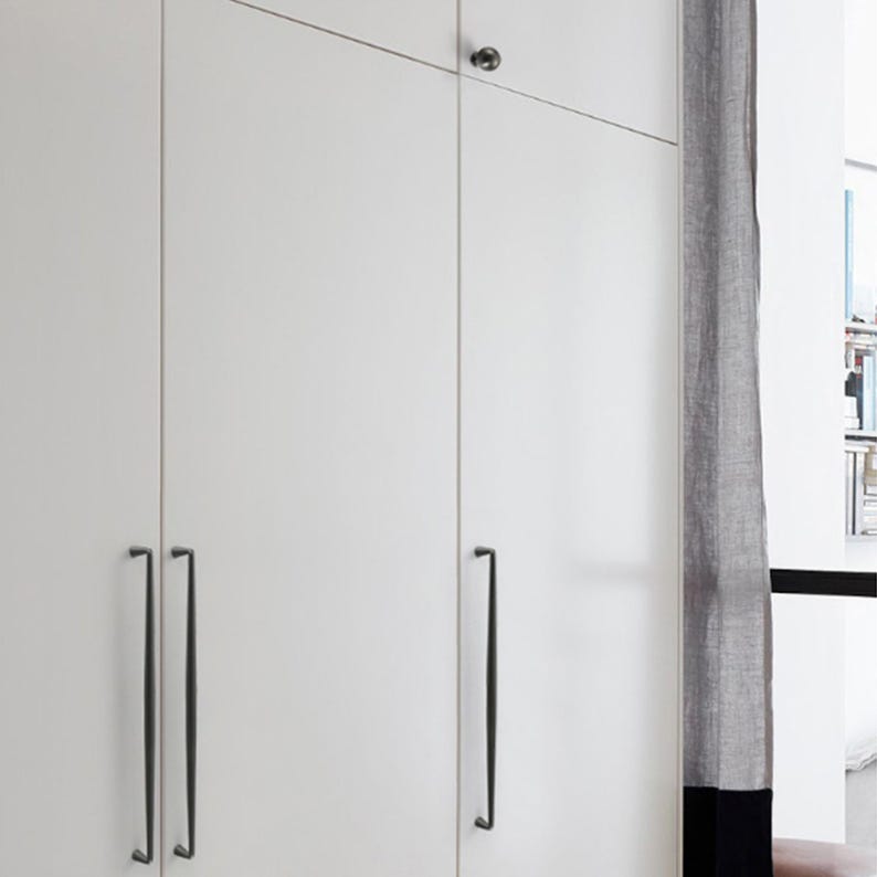 May include: A white wardrobe cabinet featuring three tall, flat-panel doors with long, slender, dark metal vertical handles and a single small, round, dark metal knob on the upper right door panel, providing a clean and functional storage solution.