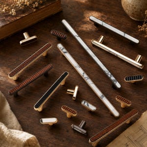 Peut inclure: Une collection de quincaillerie d'ameublement comprenant diverses poignées de tiroir et boutons. Les pièces présentent des finitions en métal doré, noir et argenté avec des inserts en marbre blanc, des textures noires striées et des motifs bois.