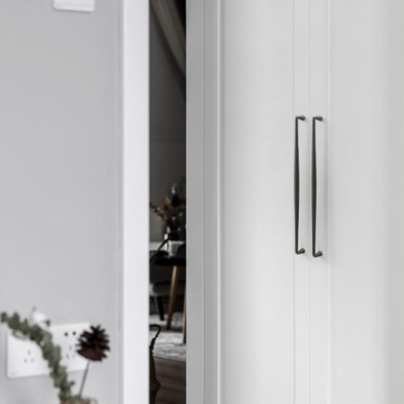 May include: A pair of long, slender, dark-toned metal door handles mounted vertically on a white paneled door. The handles feature a simple, straight design with subtle curves at the top and bottom attachment points, contrasting against the white surface.