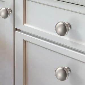 Op de afbeelding: Drie ronde kastknoppen met een geborstelde satijnnikkel afwerking. Elke knop heeft een glad, koepelvormig oppervlak en een cilindrische basis, gemonteerd op witte kastdeuren en lades met verzonken paneeldetails en strakke randen.