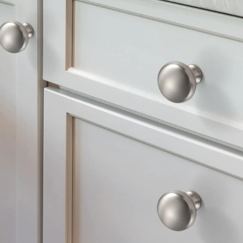 Op de afbeelding: Drie ronde kastknoppen met een geborstelde satijnnikkel afwerking. Elke knop heeft een glad, koepelvormig oppervlak en een cilindrische basis, gemonteerd op witte kastdeuren en lades met verzonken paneeldetails en strakke randen.