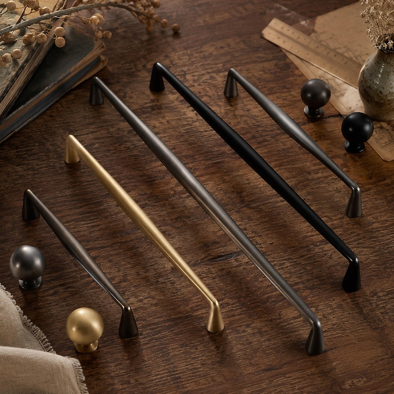 May include: A collection of metal cabinet hardware including three long, slender bar pulls in matte black, brushed gold, and dark gray finishes, accompanied by three matching round knobs in corresponding metallic colors.