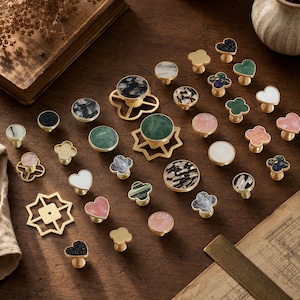 May include: A collection of decorative cabinet knobs in various shapes including circles, hearts, and clover-like designs. The knobs feature brass-toned metal bases with inlaid surfaces in marble, solid colors, and speckled patterns.
