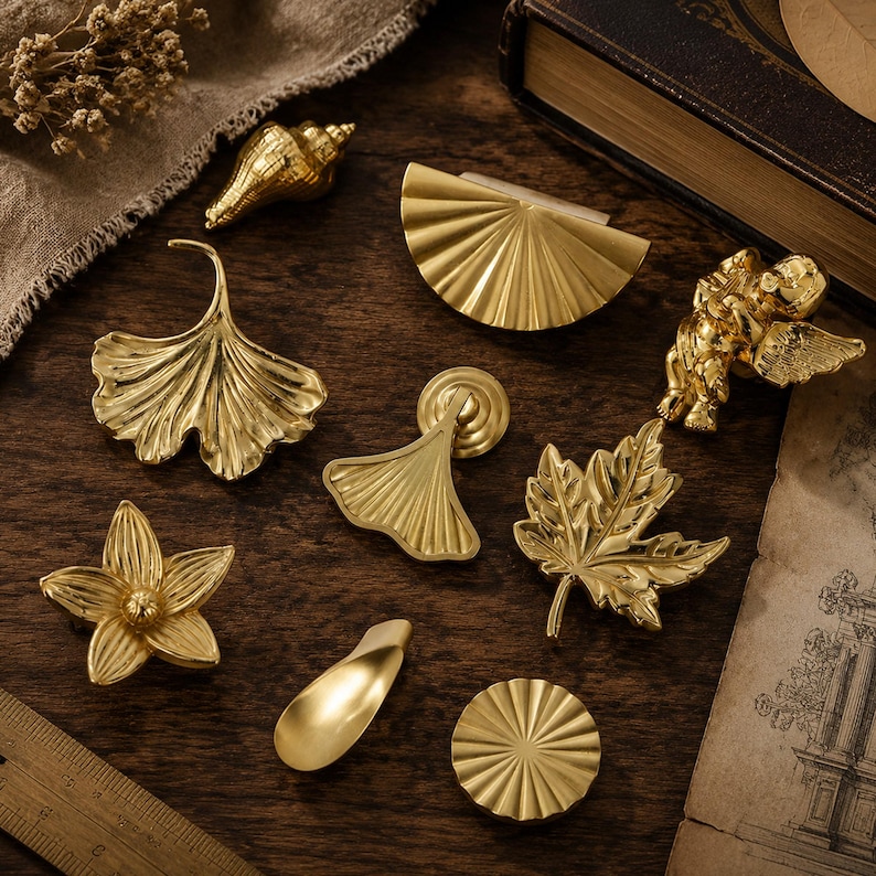 Pode incluir: Uma cole&ccedil;&atilde;o de puxadores e ma&ccedil;anetas de metal dourado em v&aacute;rias formas, incluindo uma folha de ginkgo, uma folha de bordo, uma concha, um querubim, uma flor e designs de leque plissado, sobre uma superf&iacute;cie de madeira com r&eacute;gua.