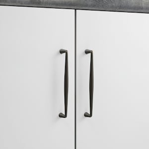 May include: Two vertical dark bronze cabinet pulls with a slender, tapered design mounted on white cabinet doors. The hardware features a smooth, matte finish and a curved profile that narrows toward the top and bottom mounting points.