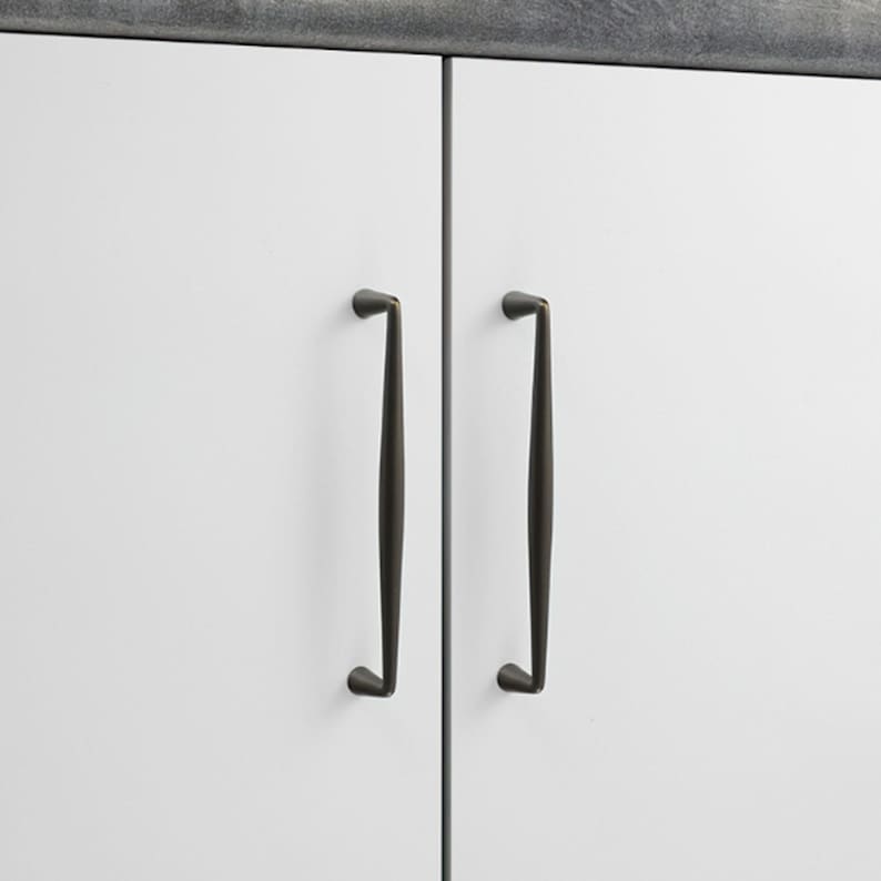 May include: Two vertical dark bronze cabinet pulls with a slender, tapered design mounted on white cabinet doors. The hardware features a smooth, matte finish and a curved profile that narrows toward the top and bottom mounting points.