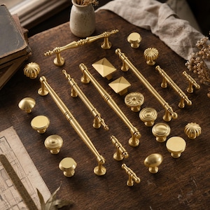 Op de afbeelding: Een collectie kastbeslag van messing, inclusief diverse ladehandgrepen, T-vormige grepen en ronde knoppen. De set bevat gladde, geribbelde en gegroefde texturen in een geborstelde gouden afwerking, gerangschikt in diverse lengtes en vormen.