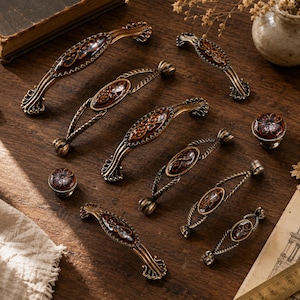 Peut inclure: Une collection de poignées et boutons de placard en métal couleur laiton antique, dotés de centres ovales avec des motifs floraux marron foncé et cuivrés. La quincaillerie comprend diverses longueurs de poignées arquées et deux boutons ronds.