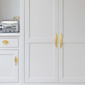 Pode incluir: Um conjunto de ferragens douradas para arm&aacute;rios, incluindo dois puxadores verticais longos em forma de pena, um puxador em forma de leque e um puxador em forma de p&aacute;ssaro instalados em arm&aacute;rios de cozinha cinza-claros com uma torradeira.