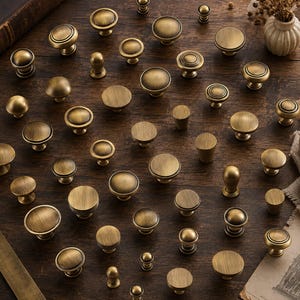 May include: A collection of various brass-toned metal cabinet knobs in different circular and cylindrical shapes, featuring brushed finishes and smooth surfaces, arranged in a scattered pattern across a dark wooden surface.
