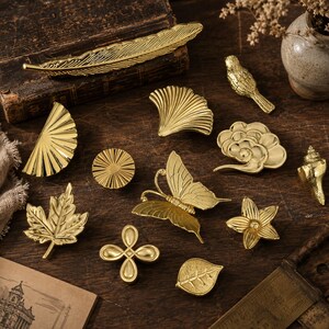 Pode incluir: Uma cole&ccedil;&atilde;o de objetos decorativos de metal dourado, incluindo uma pena, um p&aacute;ssaro, uma borboleta, uma folha de bordo, uma folha de ginkgo, uma concha, uma flor, um sol circular, um n&oacute; de quatro l&oacute;bulos e uma nuvem, todos com texturas.