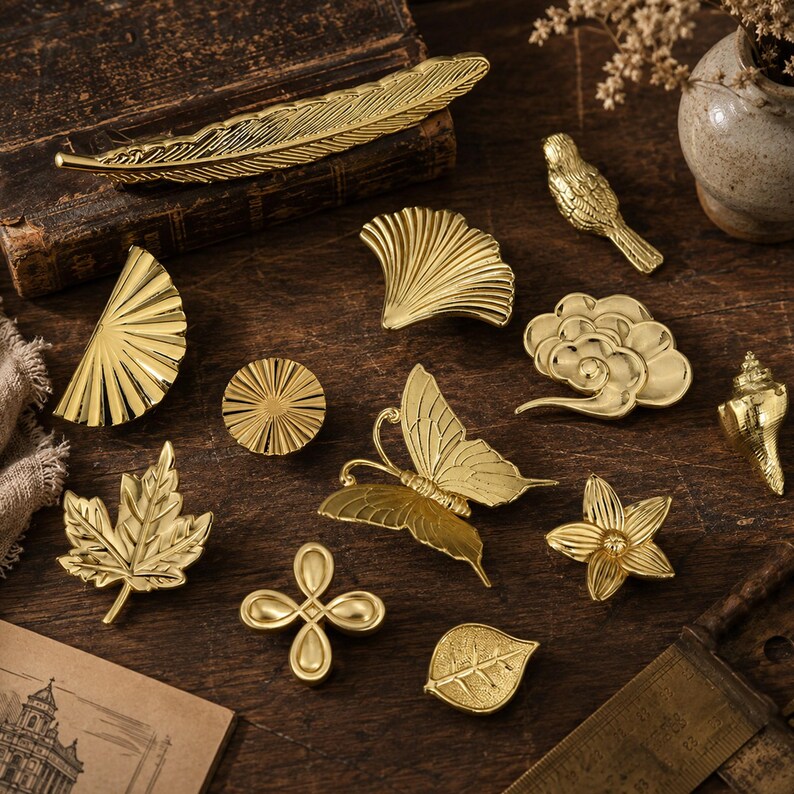 Pode incluir: Uma cole&ccedil;&atilde;o de objetos decorativos de metal dourado, incluindo uma pena, um p&aacute;ssaro, uma borboleta, uma folha de bordo, uma folha de ginkgo, uma concha, uma flor, um sol circular, um n&oacute; de quatro l&oacute;bulos e uma nuvem, todos com texturas.