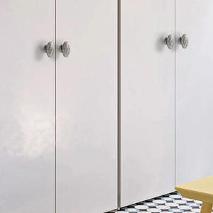 Op de afbeelding: Vier ovale, zilverkleurige metalen kastknoppen met gedetailleerde reli&euml;fpatronen zijn gemonteerd op gladde witte kastdeuren. De knoppen zijn voorzien van een centrale verticale rand en een decoratieve omlijsting voor een metalen contrast.