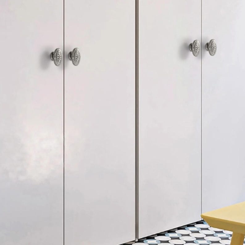 Op de afbeelding: Vier ovale, zilverkleurige metalen kastknoppen met gedetailleerde reli&euml;fpatronen zijn gemonteerd op gladde witte kastdeuren. De knoppen zijn voorzien van een centrale verticale rand en een decoratieve omlijsting voor een metalen contrast.