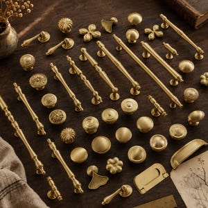 May include: A collection of brass-toned metal cabinet hardware including various drawer pulls, knobs, and handles in different shapes and sizes, arranged on a dark surface alongside a partial ruler and a piece of paper with botanical sketches.