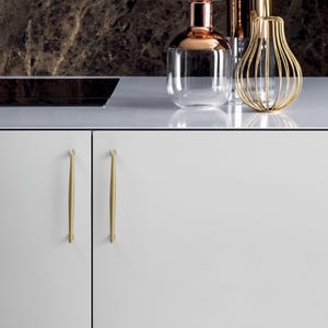 May include: Two matte gold cabinet handles with a slender, tapered design mounted on white cabinet doors. The handles feature a smooth finish and pointed ends, providing a clean look for kitchen cabinetry hardware and storage units.